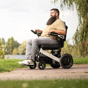 Kräftiger Mann fährt selbstständig mit dem ergoflix heavy Elektrorollstuhl auf einem Weg am Flussufer und bedient den Joystick – Seitenansicht in grüner Naturlandschaft