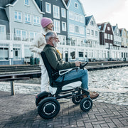 Mann fährt mit dem ergoflix vario Elektrorollstuhl entlang einer Uferpromenade mit bunten Häusern im Hintergrund und bedient den Joystick, während eine Frau hinter ihm steht und in die Ferne schaut