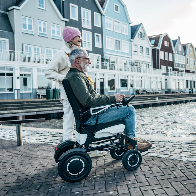 Mann fährt mit dem ergoflix vario Elektrorollstuhl entlang einer Uferpromenade mit bunten Häusern im Hintergrund und bedient den Joystick, während eine Frau hinter ihm steht und in die Ferne schaut