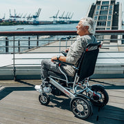 Älterer Mann fährt mit dem ergoflix basic Elektrorollstuhl auf einer Hafenpromenade und genießt den Blick auf das Wasser mit Containerkränen im Hintergrund