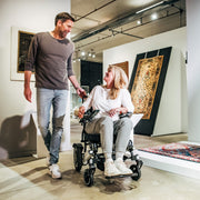 Frau fährt mit dem ergoflix LX Elektrorollstuhl durch eine Kunstgalerie und lacht mit einem Mann, der neben ihr herläuft – moderne Ausstellungsräume im Hintergrund