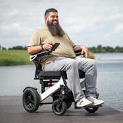 Lächelnder kräftiger Mann sitzt in dem ergoflix heavy Elektrorollstuhl auf einem Steg am See und hält sein Smartphone in der Hand – Frontansicht im Freien