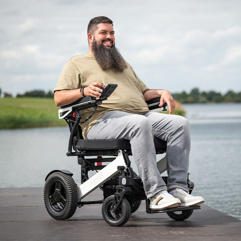 Lächelnder kräftiger Mann sitzt in dem ergoflix heavy Elektrorollstuhl auf einem Steg am See und hält sein Smartphone in der Hand – Frontansicht im Freien