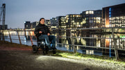 Mann fährt abends mit dem ergoflix carbon Elektrorollstuhl mit eingeschaltetem Frontlicht an einer Uferpromenade entlang – im Hintergrund spiegeln sich beleuchtete Stadtgebäude im Wasser