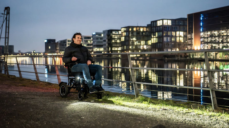 Mann fährt abends mit dem ergoflix carbon Elektrorollstuhl mit eingeschaltetem Frontlicht an einer Uferpromenade entlang – im Hintergrund spiegeln sich beleuchtete Stadtgebäude im Wasser